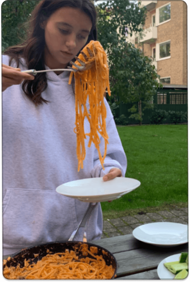 Una persona joven sostiene un plato de fideos junto a una mesa con comida, que incluye una bandeja de fideos y verduras en rodajas, en un área exterior residencial.