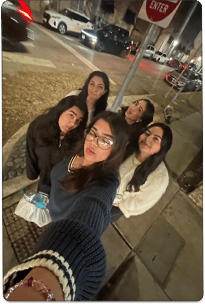 Un grupo de jóvenes posa junto en una acera por la noche, sonriendo hacia la cámara, con autos estacionados y señales de tránsito al fondo.