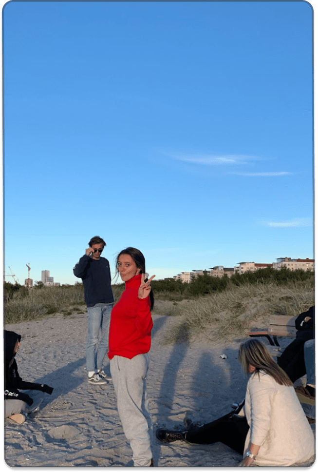 Un grupo de jóvenes pasa tiempo junto en una zona de playa o dunas de arena bajo un cielo azul despejado, con varias personas de pie y sentadas al fondo.